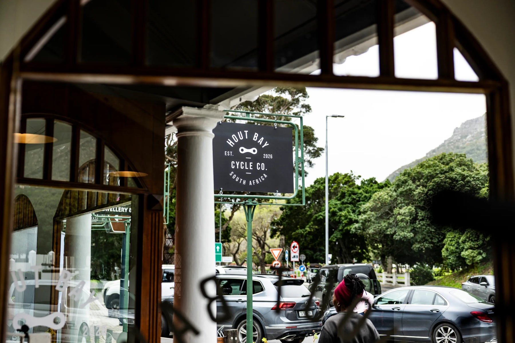 The Hout Bay Cycle Co sign on Main Road, seen from inside the shop, with the mountain behind